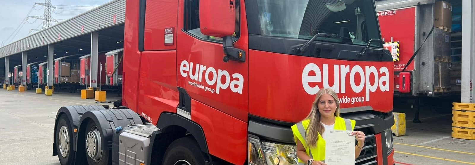 Darcy Cook stands in front of a red branded Europa truck in a Hi Vis holding here CPC certificate
