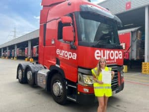 Darcy Cook stands in front of a red branded Europa truck in a Hi Vis holding here CPC certificate