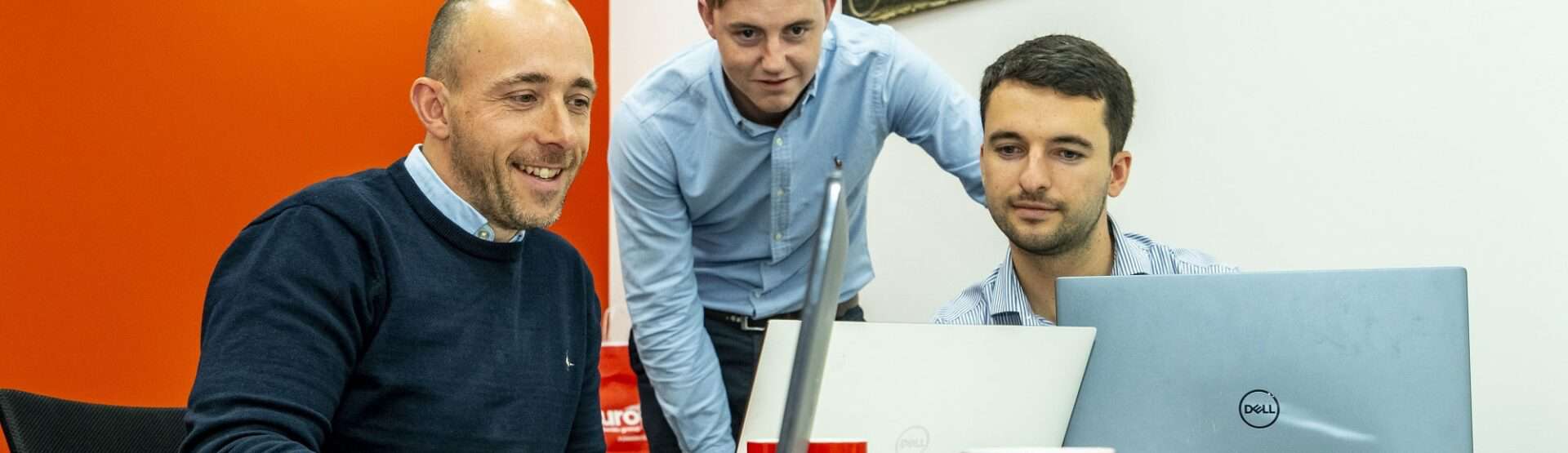Adrian, Jamie and Elliott sit round a laptop that they're all looking at. A red and white wall are in the background